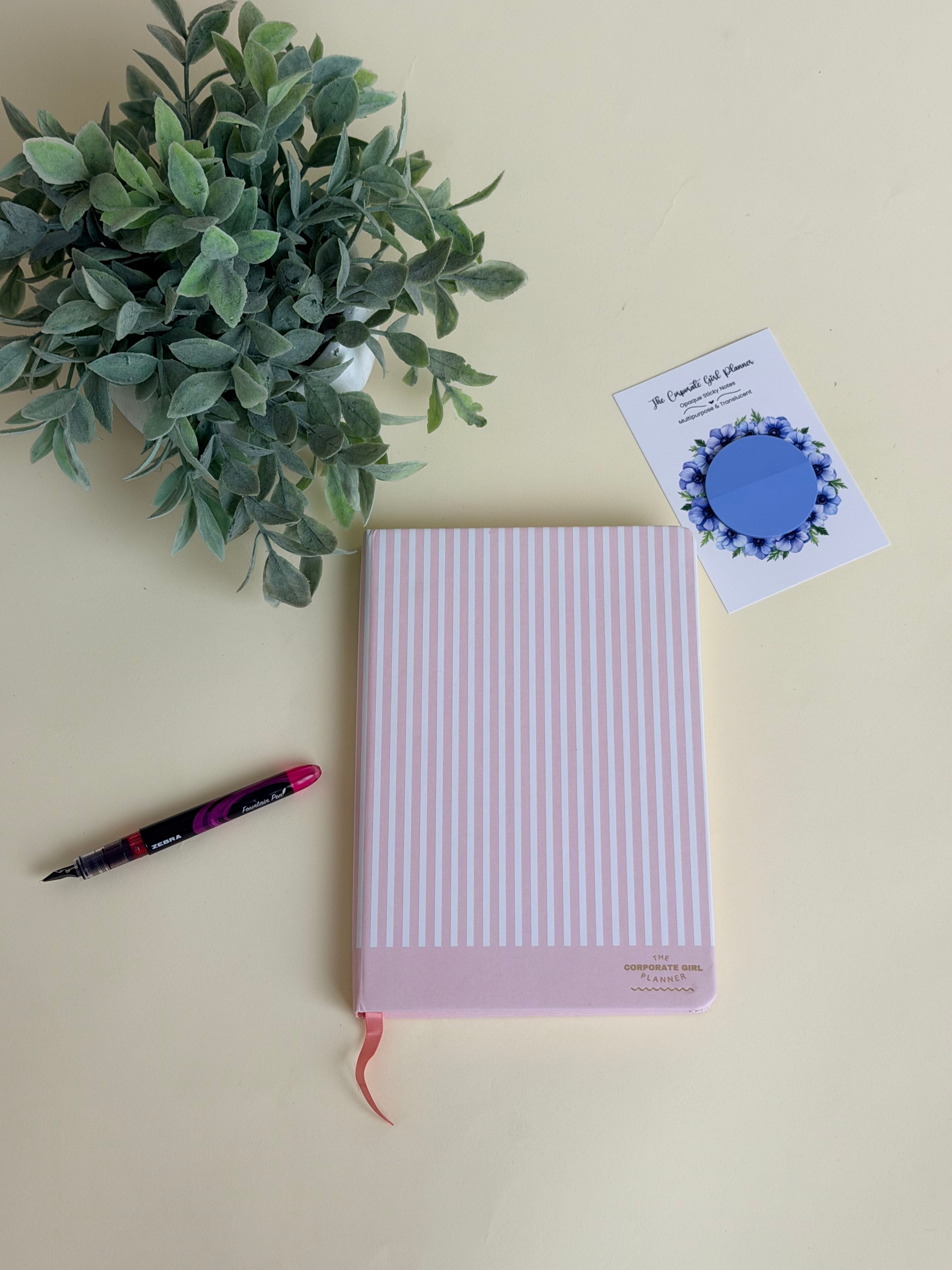 A pink and white vertically striped hardcover notebook with a pink ribbon bookmark, styled on a cream background with a green plant, a black and pink pen, and a Corporate Girl Sticky Notes.