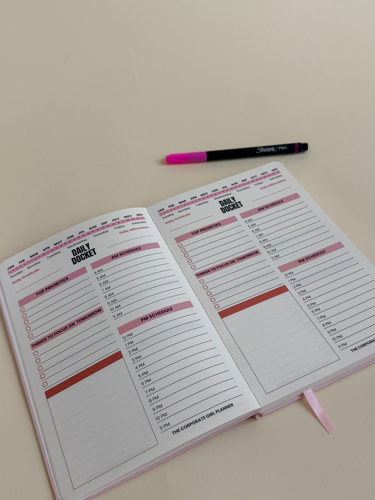 An open planner or notebook showing printed daily Docket pages with lined sections and pink headers, placed on a light beige surface next to a black and pink pen.