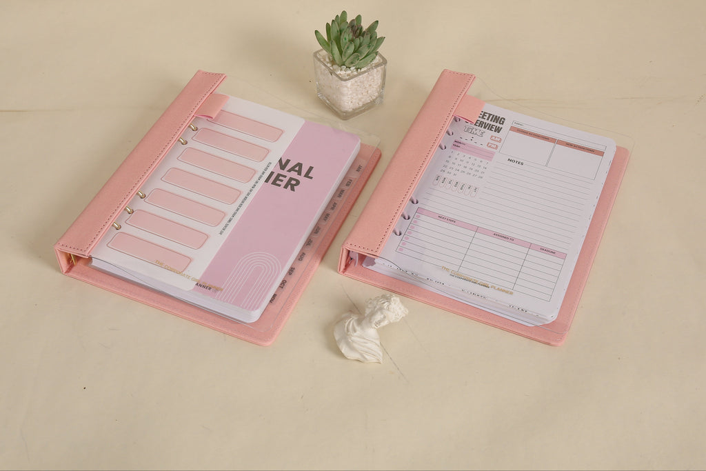 Two pink planners open on a beige surface with a small plant in the background.