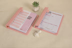 Two pink planners open on a beige surface with a small plant in the background.