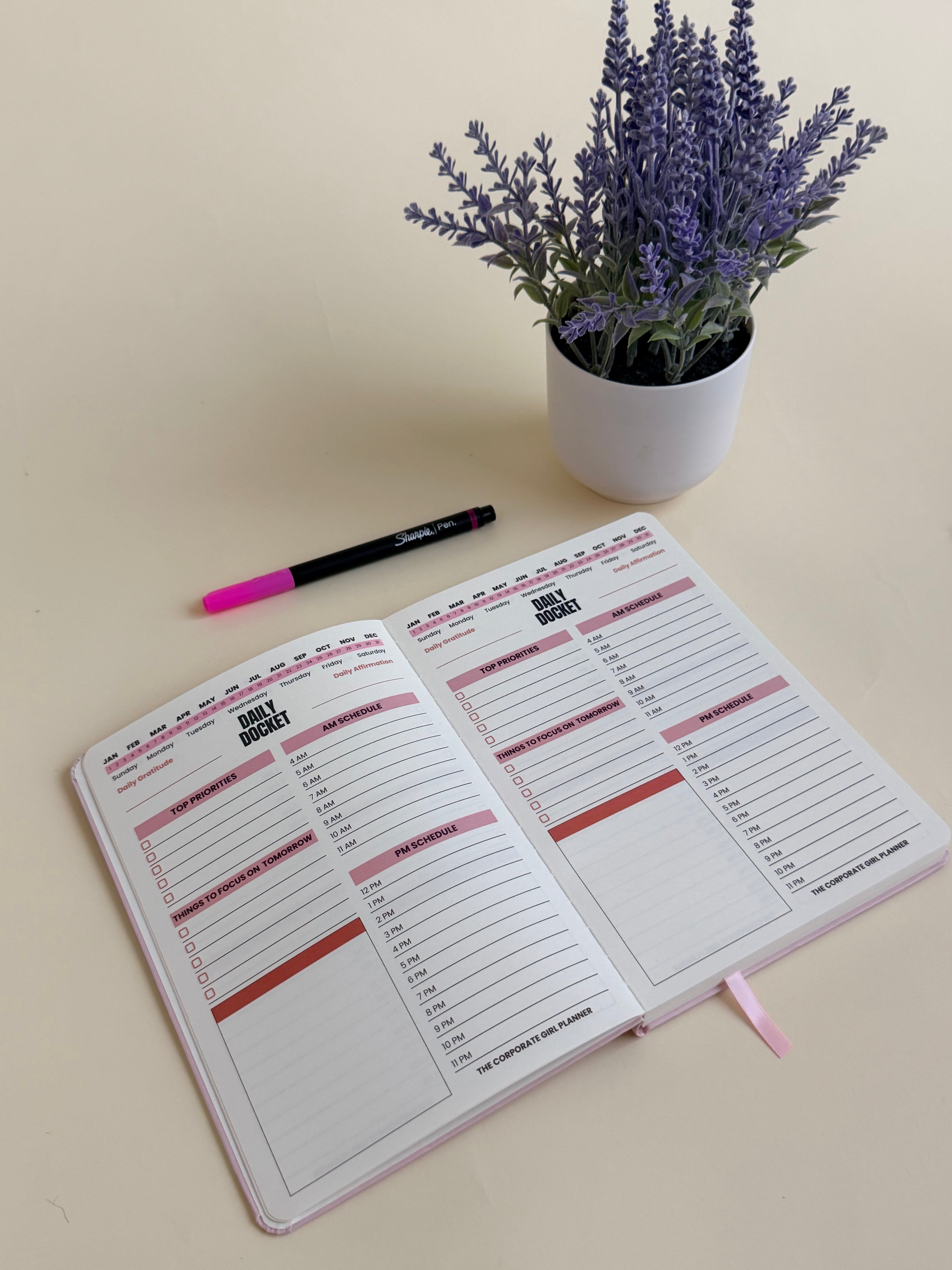 An open planner, notebook and agenda with lined Daily Docket pages and pink accents, styled on a light beige surface next to a small white pot of lavender and a black pen with a pink cap.